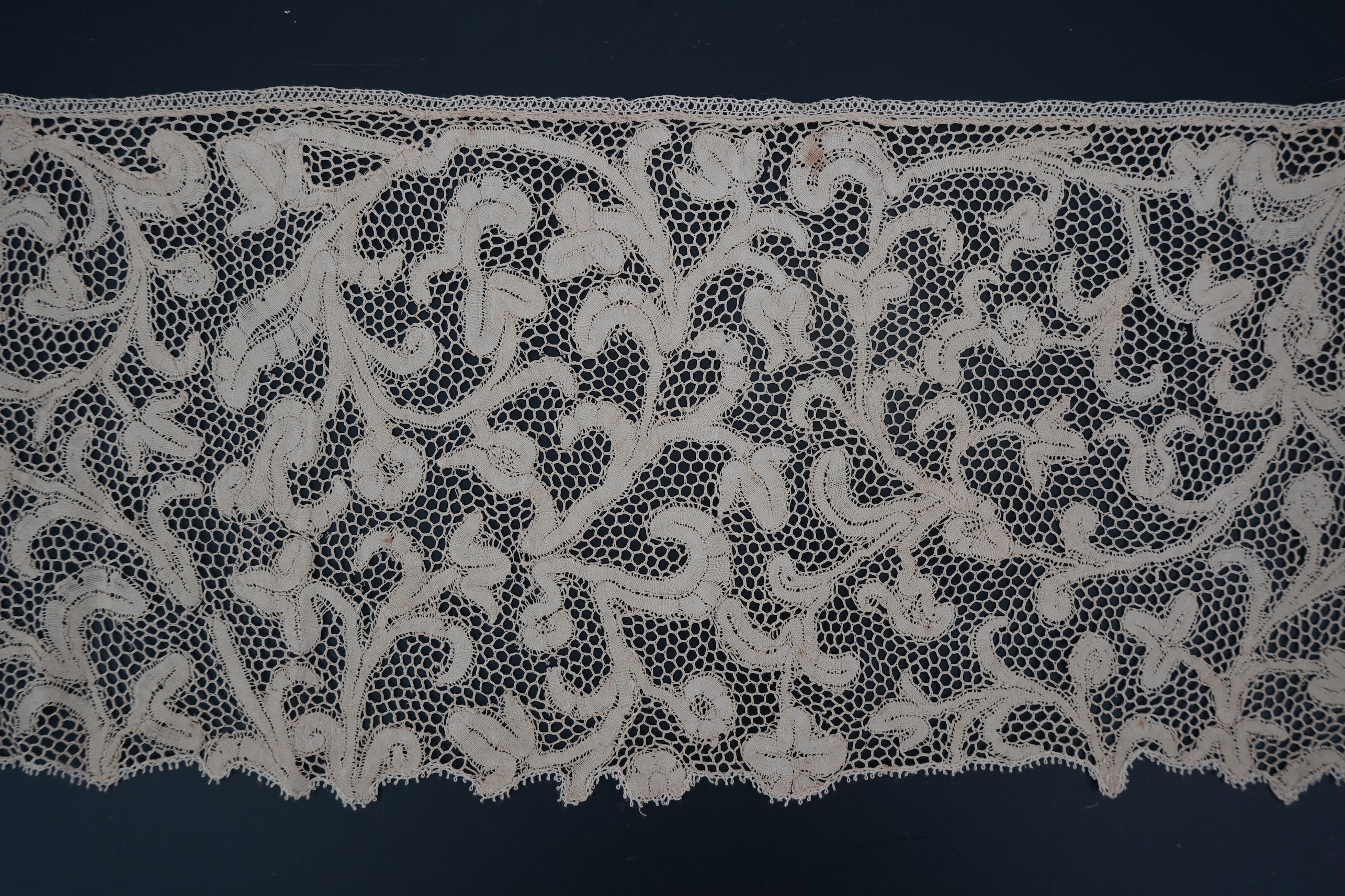 A length of 1740-1760 Italian, Milanese bobbin lace flounce, made from flat braiding or tapes, placed into scrolling patterns, attached to brides, made into a honeycomb mesh. 252cm long x 16cm deep. Condition - one small